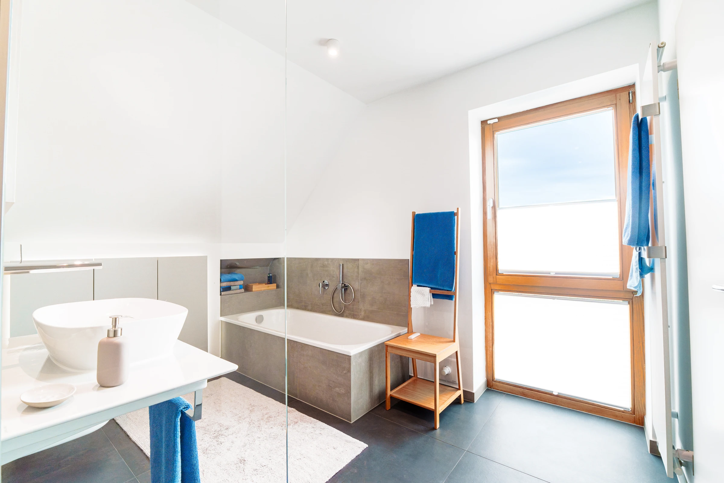 Lichtdurchflutetes Bad mit Wanne, Glasdusche, graubraunen Fliesen und blauen Handtüchern
