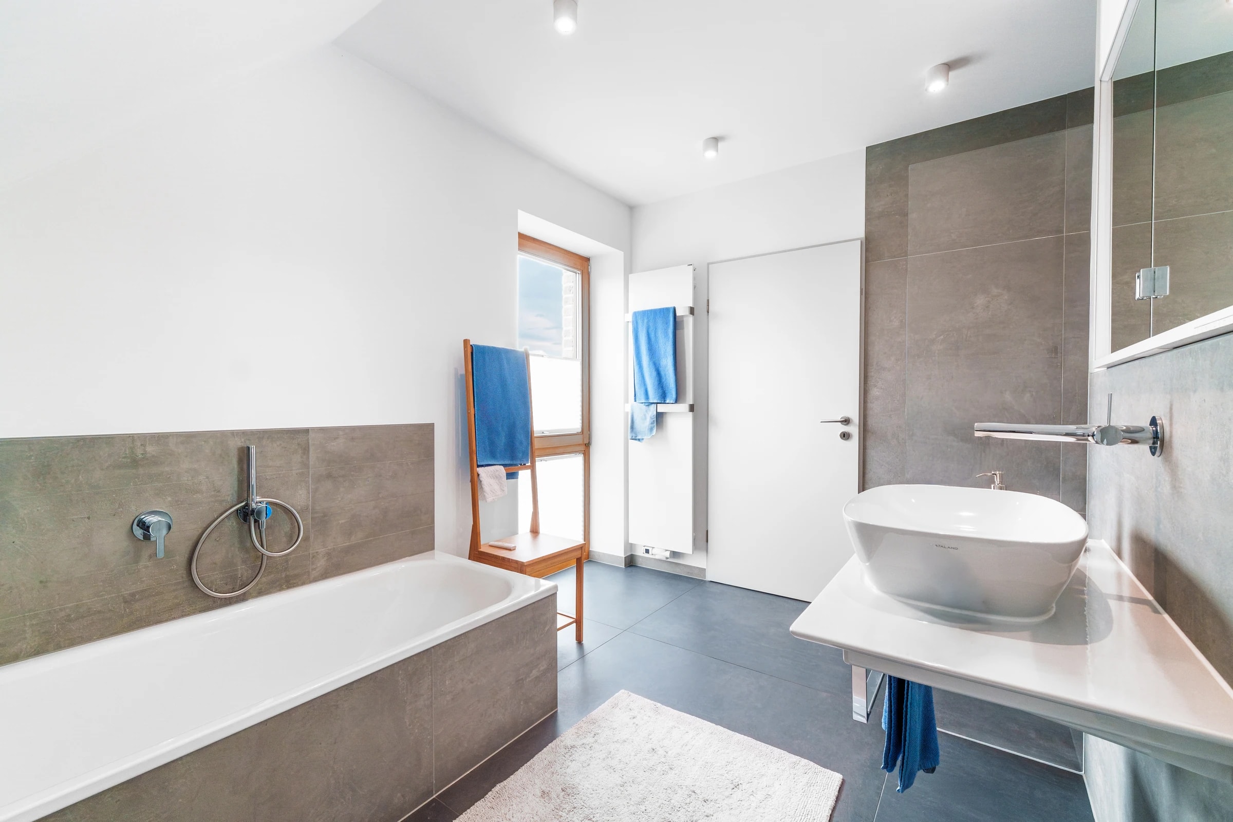 Lichtdurchflutetes Bad mit Wanne, Glasdusche, graubraunen Fliesen und blauen Handtüchern